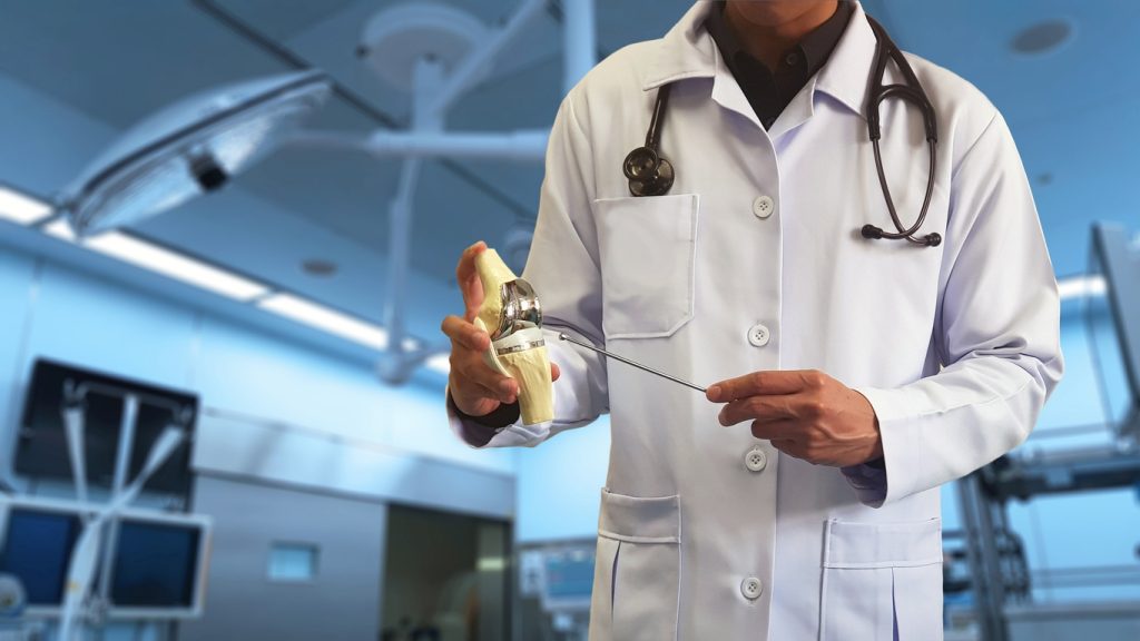 Orthopedic Doctor Showing Model Of Total Knee Replacement(tkr) With Blurred Background Of Operative Bed In Modern Operating Room. Medical Technology And Operative Care Concept.